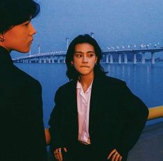 two people standing next to each other in front of a body of water with a bridge in the background