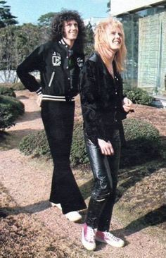 two people standing next to each other in front of bushes and shrubbery, with one person wearing black leather pants