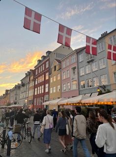 many people are walking on the sidewalk near some buildings and flags flying in the air