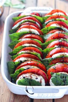 a white dish filled with sliced tomatoes covered in cheese and herbs on top of a wooden table