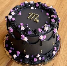 a black cake with purple flowers and the letter m on top is sitting on a wooden table