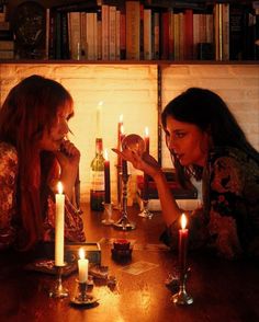 two women sitting at a table with candles in front of them and books on the shelves behind them