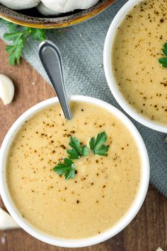 two white bowls filled with soup on top of a wooden table next to garlic and parsley