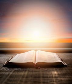 an open book sitting on top of a wooden table next to the ocean at sunset