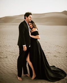 a man and woman standing in the sand with their arms around each other, posing for a photo