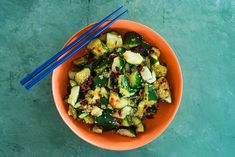 an orange bowl filled with cucumbers and other vegetables next to two blue chopsticks