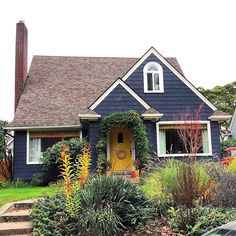 a blue house with yellow door surrounded by greenery and flowers on the front lawn