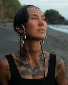 a woman with tattoos and piercings on her head standing in front of the ocean