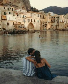 a man and woman sitting next to each other on the edge of a body of water