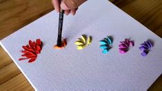 a person holding a paintbrush in front of four different colored paper flowers on a table