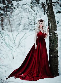 a woman in a long red dress standing next to a tree with snow on it