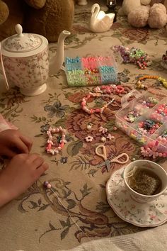 a table topped with lots of different types of beads and cup of tea next to a teddy bear