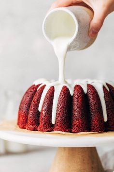 someone pouring icing onto a red velvet bundt cake on a white platter