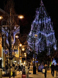 people are walking down the street in front of a christmas tree with lights on it