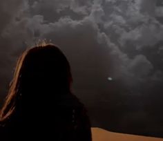 a woman standing in front of a cloudy sky with the sun shining through her hair