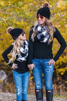 two women standing next to each other wearing black boots and scarfs, with trees in the background