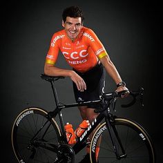 a man in an orange shirt and black shorts poses with his bike against a dark background