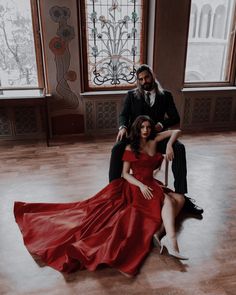 a man and woman sitting on the floor in front of two windows wearing red dresses