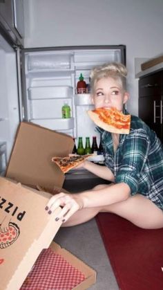 a woman sitting on the floor eating a slice of pizza in front of an open refrigerator