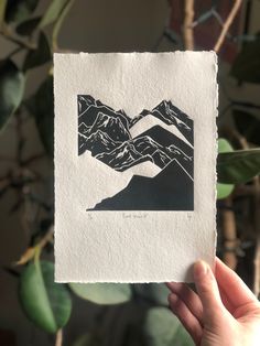 a hand holding up a piece of paper with mountains on it in front of a plant