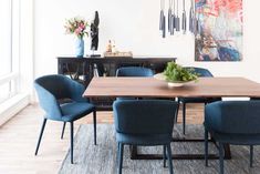 a dining room table with blue chairs and a vase on top of it in front of a painting