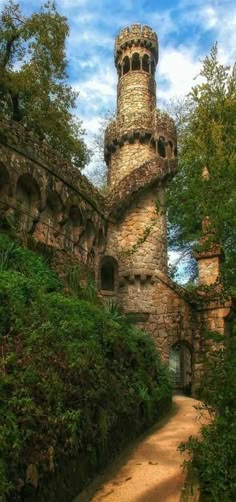 an old stone building with a tower on top surrounded by greenery and trees in front of a cloudy blue sky