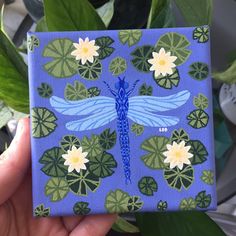 a blue dragonfly painted on a purple background surrounded by lily pads and water lilies