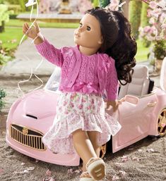 a doll is sitting in front of a pink car and holding a string with her hand