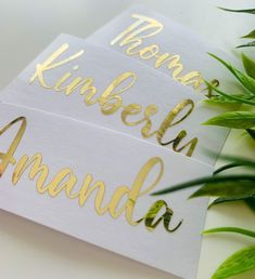 two white cards with gold lettering on them next to some green plants and leaves in the background