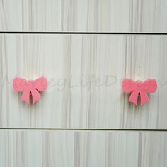 two pieces of pink paper with bows attached to the side of a white wooden wall