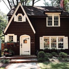 a brown house with white trim and two windows on the front door is surrounded by greenery