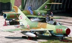 two green fighter jets parked next to each other on top of a brick floored area