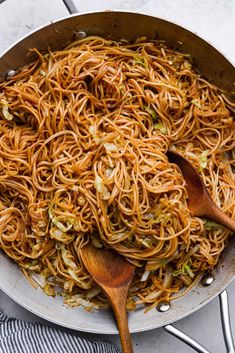 a skillet filled with noodles and vegetables on top of a white tablecloth next to two wooden spoons