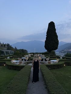 a woman in a long black dress walking down a path through a formal garden area