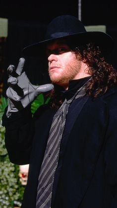 a man with long hair wearing a black hat and tie holding a fake hand up to his face