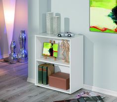 a white book shelf filled with books next to a painting and vases on the wall