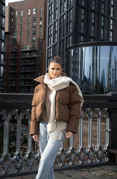 Woman wearing a brown puffer jacket paired with a cream scarf and light-wash jeans, standing outdoors against a backdrop of modern city buildings for a stylish urban winter look. Brown Down Jacket Outfit, Brown Puffer Jacket Outfit Winter, Khaki Puffer Coat Outfit, Brown Puff Jacket Outfit, Brown Jacket Winter Outfit, Brown Leather Puffer Jacket Outfit, Light Brown Puffer Jacket Outfit, Brown Winter Jacket Outfit, Cropped Puffer Coat Outfit