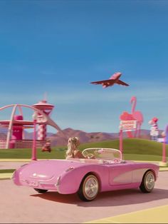 a pink car driving down a street next to a park with an airplane in the sky