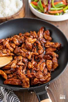 the chicken is being cooked in a skillet with a wooden spoon and rice on the side