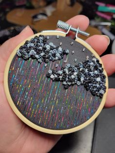 a hand is holding a small embroidery hoop with black beads and silver thread on it