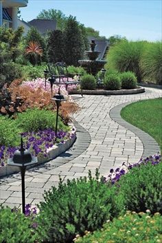 a garden with flowers and plants around it in the middle of a sidewalk path surrounded by greenery