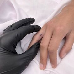 a person's hand with a small cross tattoo on their ring finger, resting on a white sheet