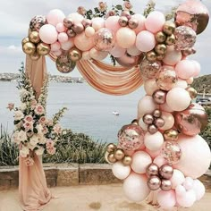 a wedding arch decorated with pink and gold balloons, flowers and greenery on the beach