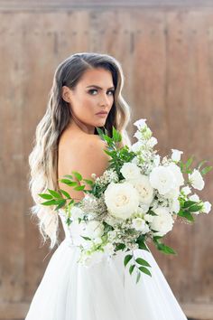 a woman in a wedding dress holding a bouquet of white flowers and greenery with her back to the camera