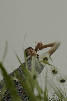 a woman standing in tall grass with her hand up to her face and looking up at the sky