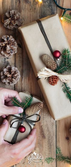 a person holding a wrapped present box with pine cones and ornaments around it on a wooden table