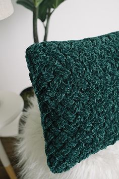 a green crocheted pillow sitting on top of a white fur covered floor next to a potted plant