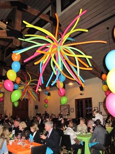 a large group of people sitting at tables in a room with balloons hanging from the ceiling