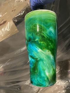 a hand holding a glass vase with green and blue swirls on it, in front of plastic bags