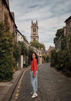 a woman standing in the middle of an alley with buildings on both sides and a clock tower behind her